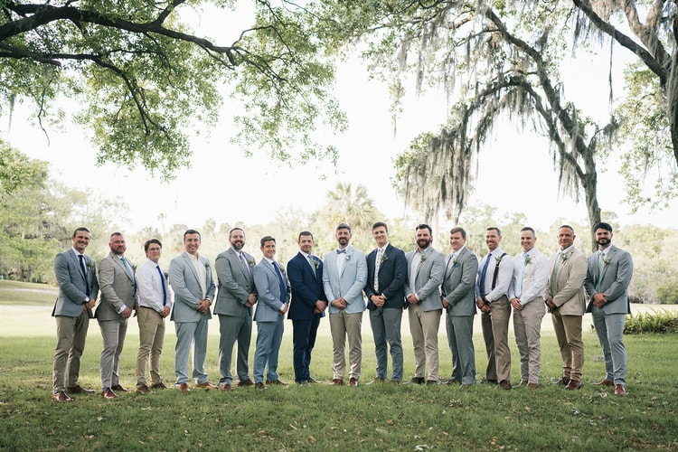 Large wedding party: groom stands at center flanked by groomsmen in khaki, tan and navy suits on a sun-lit lawn, wedding photography
