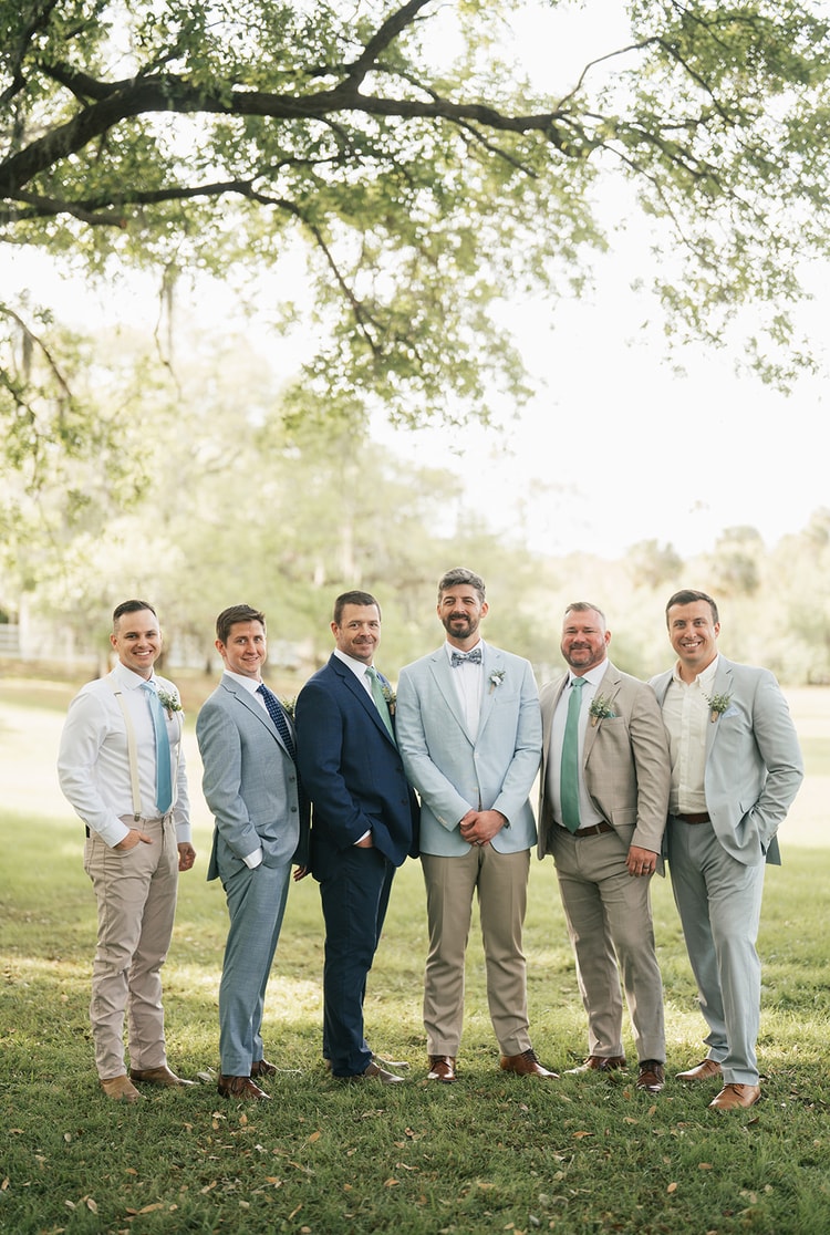 Groom in a light blue suit and floral bow tie stands with five groomsmen in mixed blue and tan suits on a grassy lawn, wedding photography