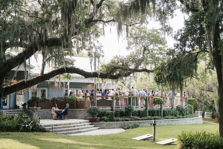 Guests mingle on a deck terrace under sweeping oak branches, a lawn with cornhole in the foreground, wedding photography