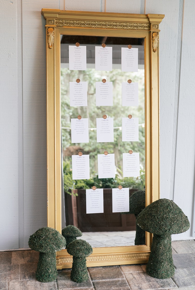Seating chart cards pinned to a gold ornate mirror, flanked by moss mushroom sculptures at the reception entrance, wedding details