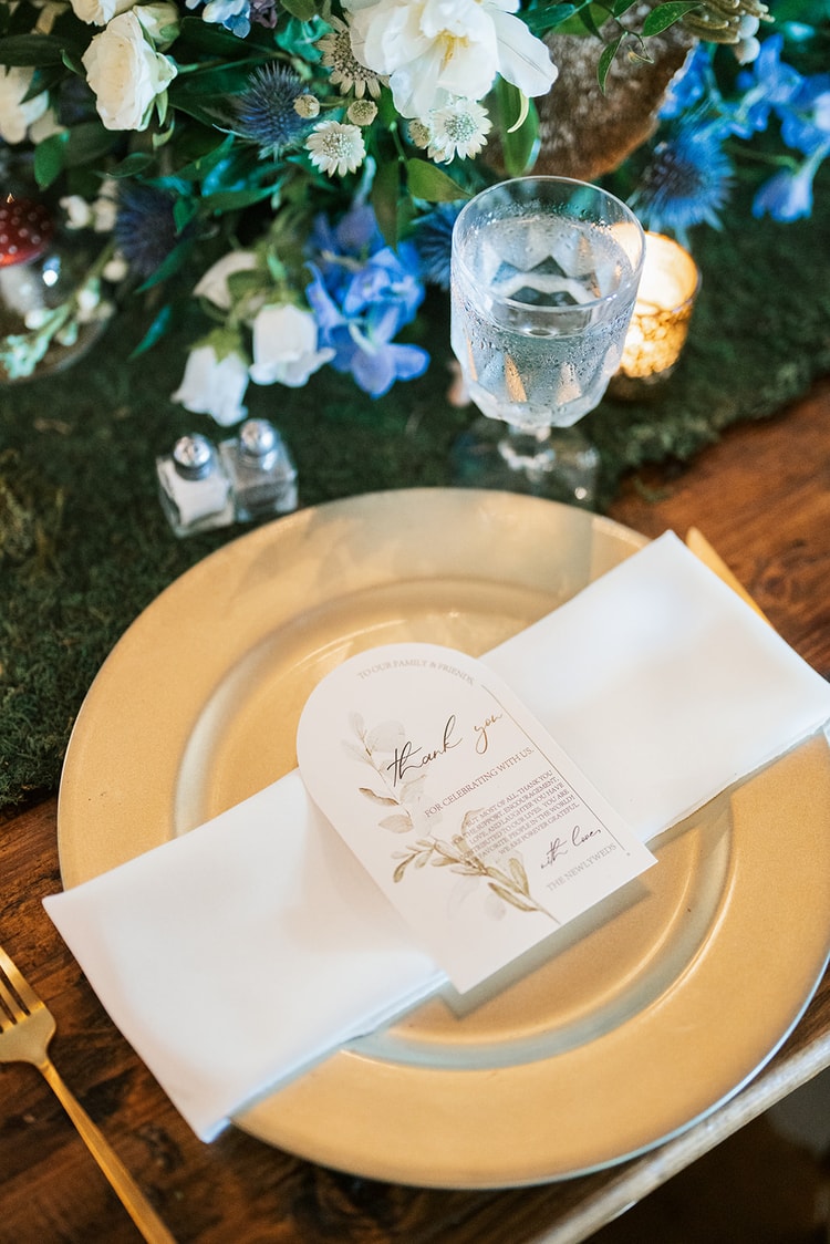 Place setting detail: gold charger plate with white folded napkin and an arched thank-you card, crystal glassware and blue floral centerpiece above, wedding details