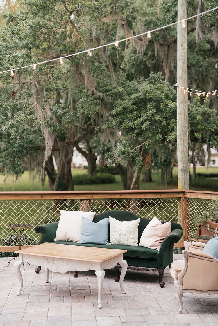 Outdoor cocktail lounge: a dark green velvet sofa with pastel cushions faces a white carved coffee table under string lights and mossy oaks, wedding details