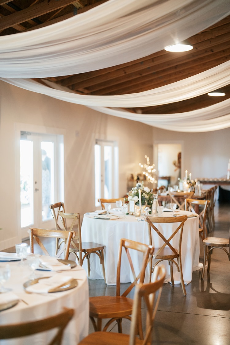 Reception hall with white fabric-draped ceiling beams, round tables set with white linens and cross-back wooden chairs, warm candlelight glowing, wedding details