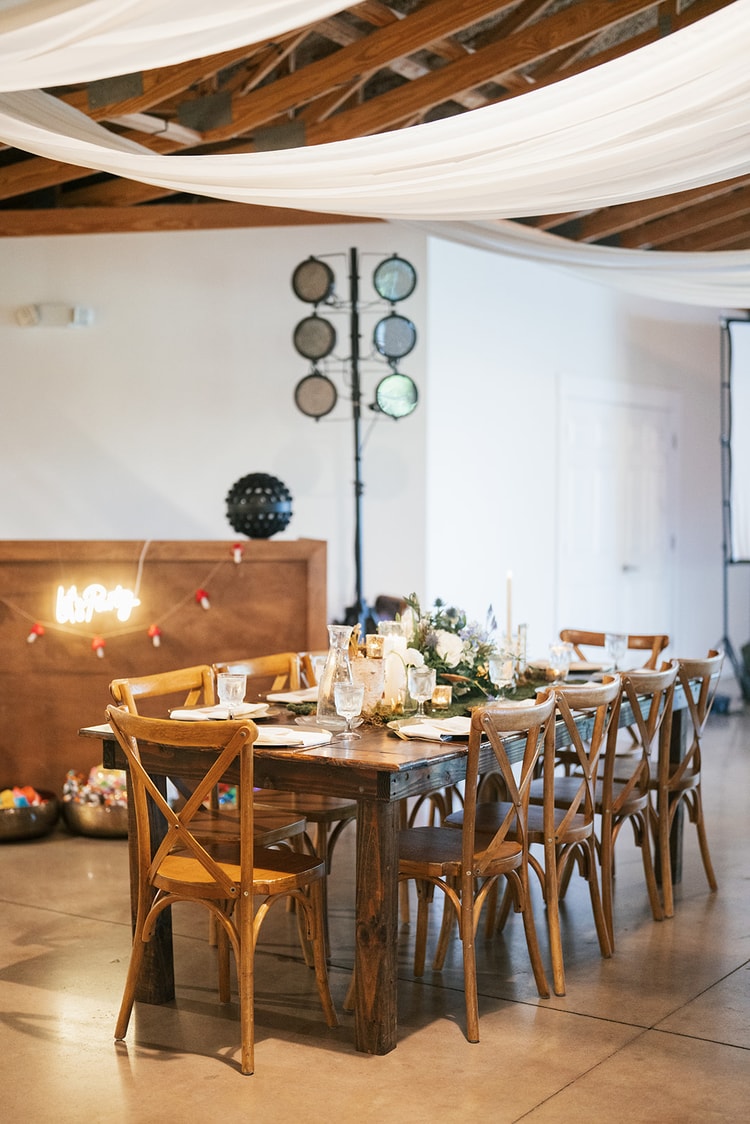 Rustic barn reception hall set for dinner, farm tables with cross-back chairs under white fabric ceiling draping and a neon sign glowing on the wall, wedding details