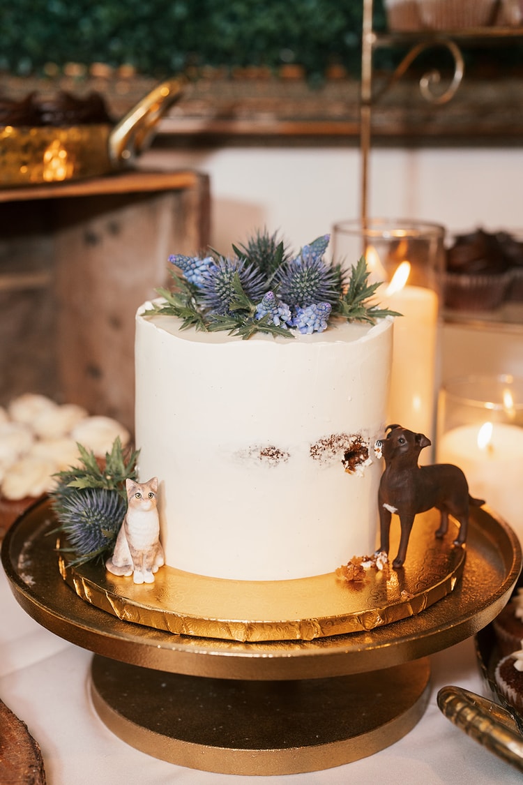 White single-tier wedding cake topped with blue thistles and muscari flowers, adorned with a cat and dog figurine on a gold stand, wedding details