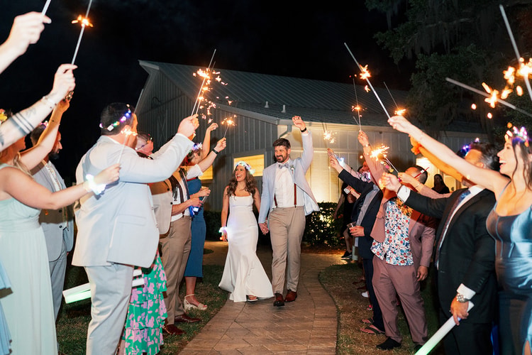 Bride and groom walk hand-in-hand through a sparkler exit tunnel surrounded by cheering guests at night