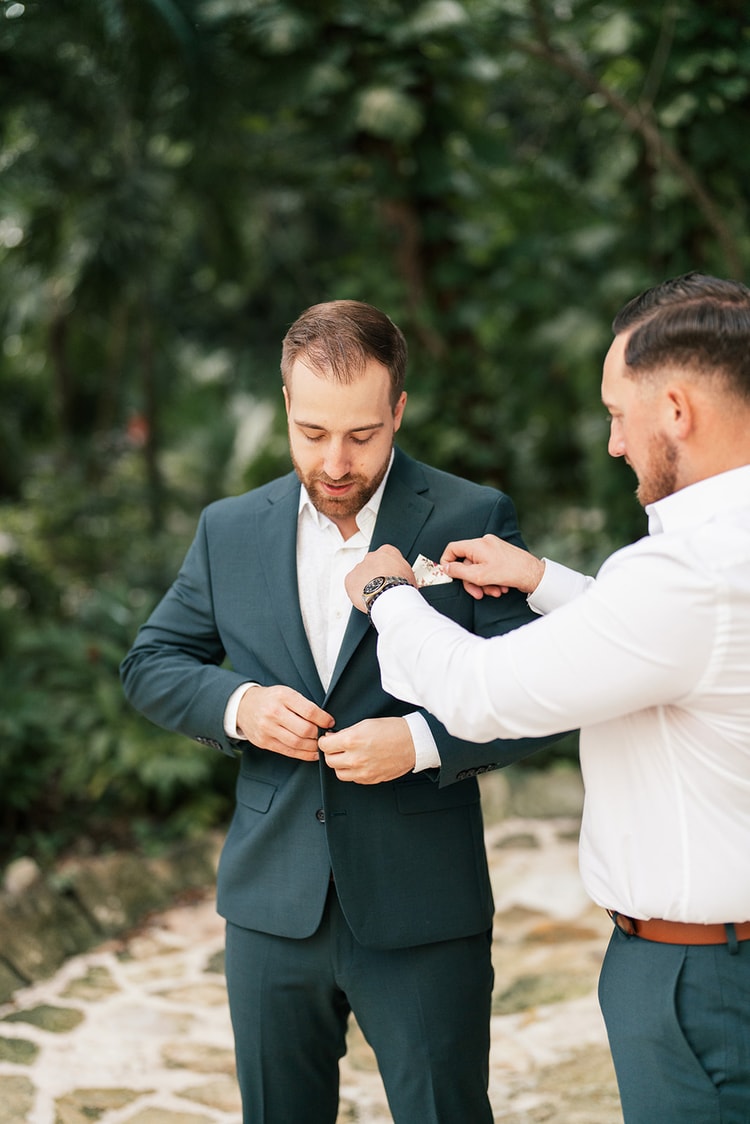 Groomsman tucking a floral pocket square into the groom's dark teal suit jacket while getting ready on a stone garden path