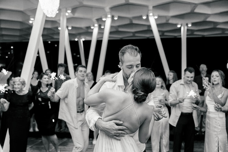 Bride and groom embrace and kiss on the dance floor as guests wave sparklers around them at night.