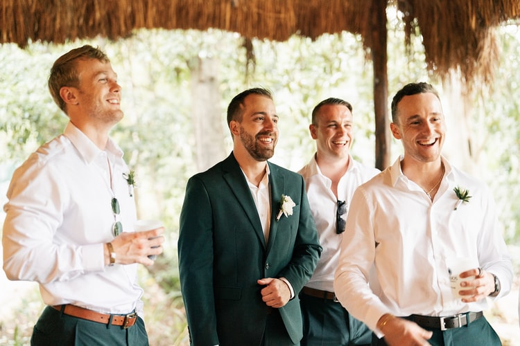 Groom in a green suit laughs with three groomsmen holding drinks under a palapa roof before the ceremony.