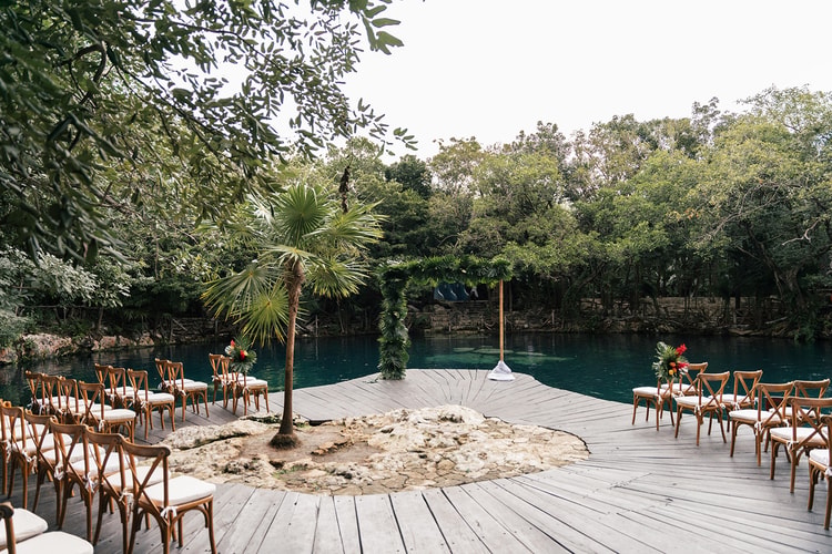 Empty wooden ceremony chairs with white cushions line a dock beside a turquoise cenote, a palm-covered arch waiting at the water's edge in Playa del Carmen.