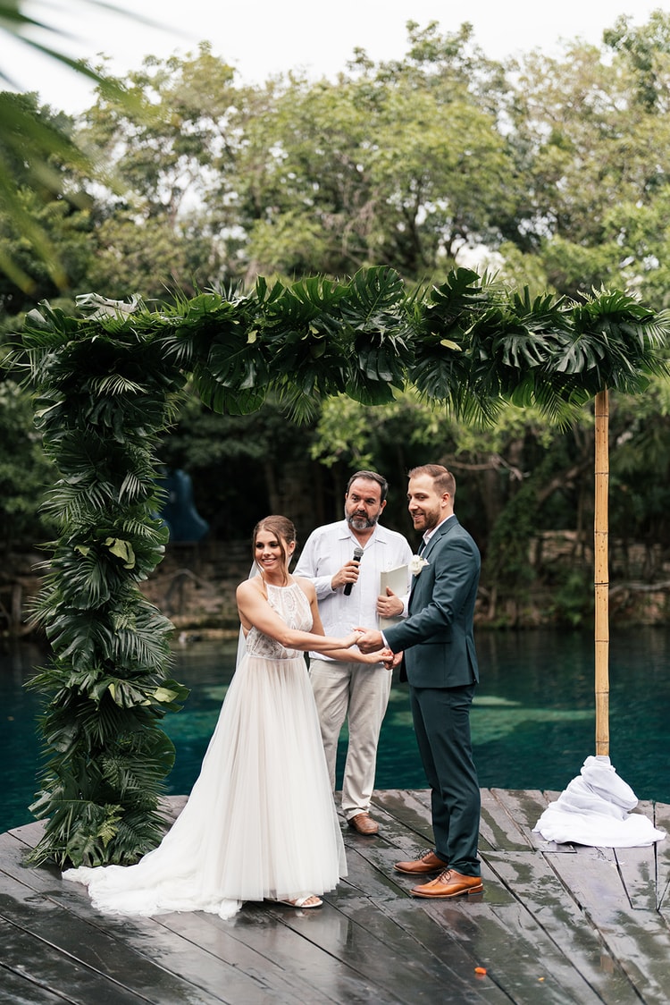 Bride and groom hold hands and smile as the officiant reads from a book beneath a palm-leaf ceremony arch over cenote water.