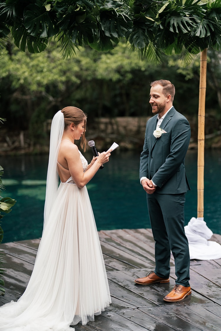 Bride in a backless lace gown reads her vows aloud to the groom under a monstera-leaf arch beside turquoise water.