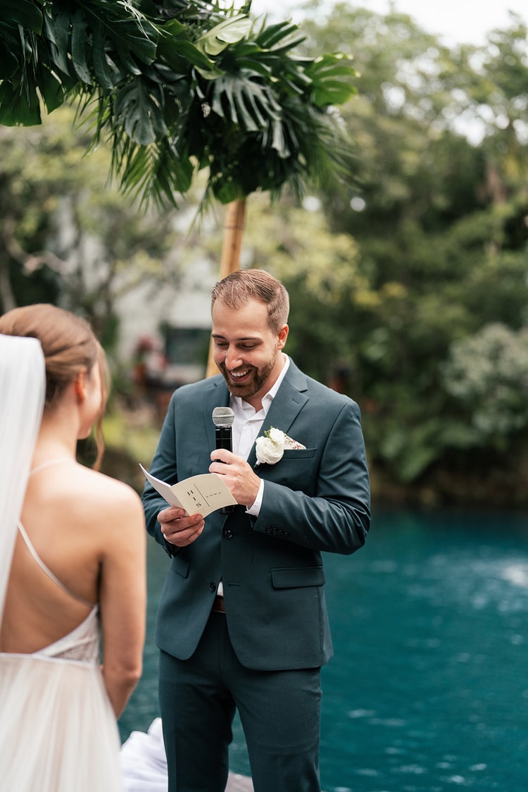 Groom in a teal suit smiles while reading his vows from a card into a microphone beneath a tropical leaf arch.