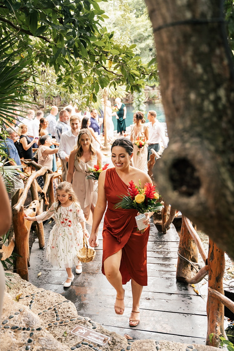 Bridesmaid in a rust dress and a young flower girl carrying a basket walk down a wooden boardwalk past smiling guests.