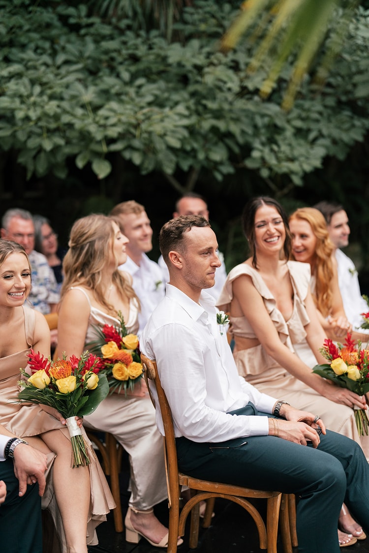 Groom in a white shirt sits among laughing bridesmaids and groomsmen holding bright bouquets during the ceremony.