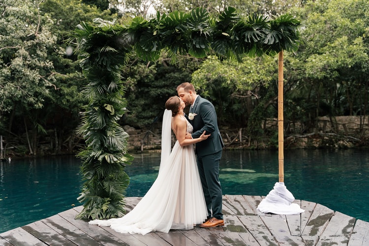 Bride and groom share a first kiss under a tropical leaf-draped arch on a dock beside a cenote in Playa del Carmen.
