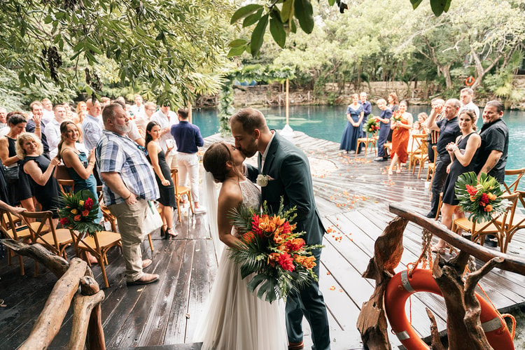 Newlyweds kiss while walking past cheering guests and lifebuoys along a wooden dock beside turquoise cenote water.
