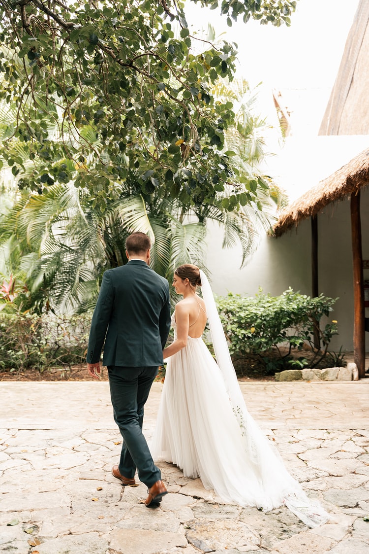 Bride and groom hold hands and walk away together past palm trees toward a thatched-roof building.