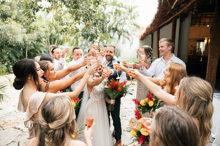 Wedding party raises champagne glasses in a toast around the bride and groom outside a palapa-roofed reception space.
