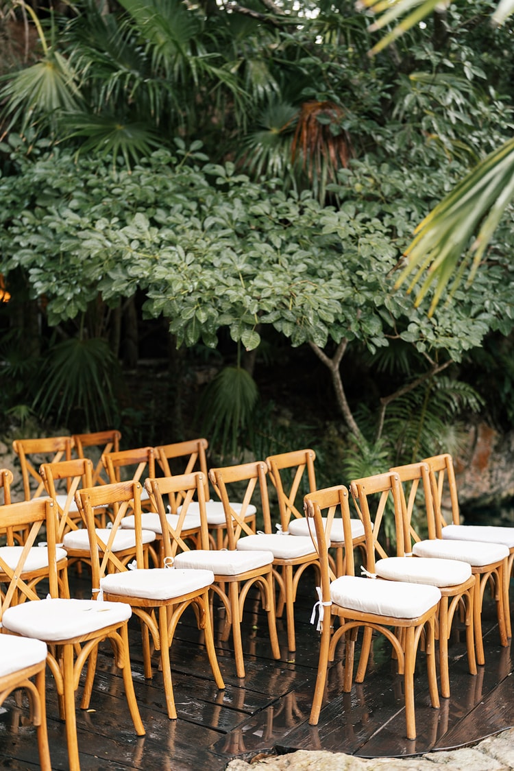 Empty cross-back ceremony chairs on a wet dock with the grey light and jungle tree line reflected in the water