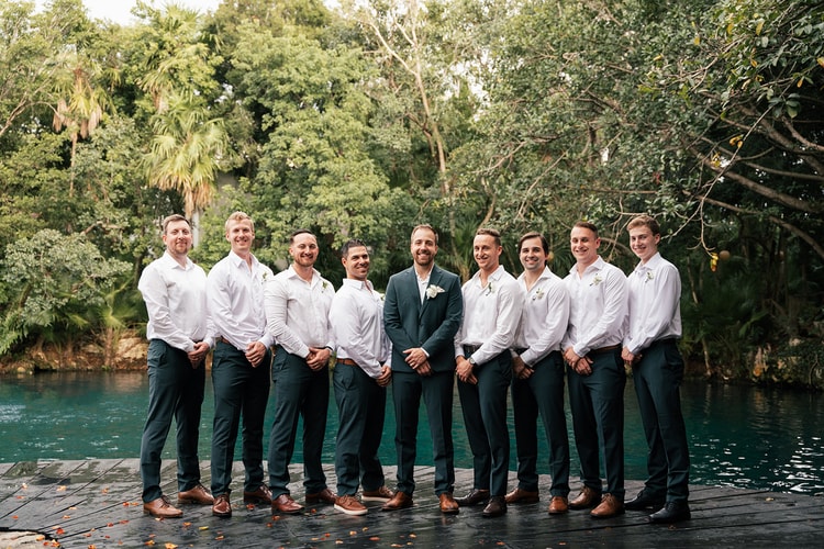 Groomsmen in dark green suits and white shirts line up on a dock beside teal water, smiling at the camera.