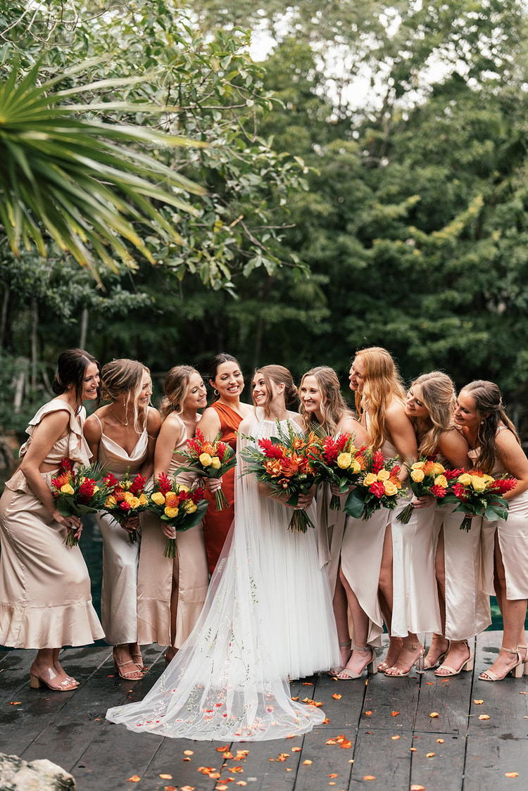Bride and her bridesmaids huddle close together on a dock, laughing and holding tropical bouquets by a jungle lagoon.