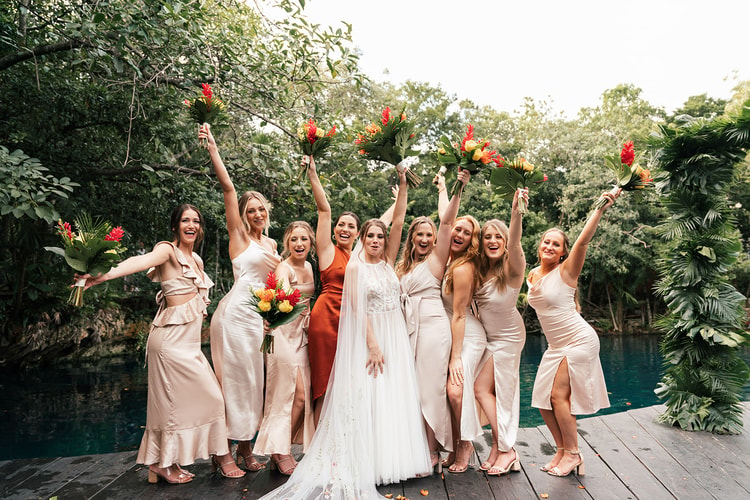 Bride and bridesmaids raise their bouquets and cheer together on a dock by a lagoon in Playa del Carmen.