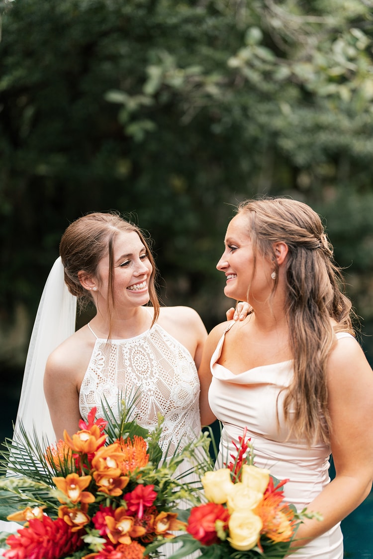 Bride and a bridesmaid in a blush dress smile at each other, holding tropical bouquets of orange and red blooms.