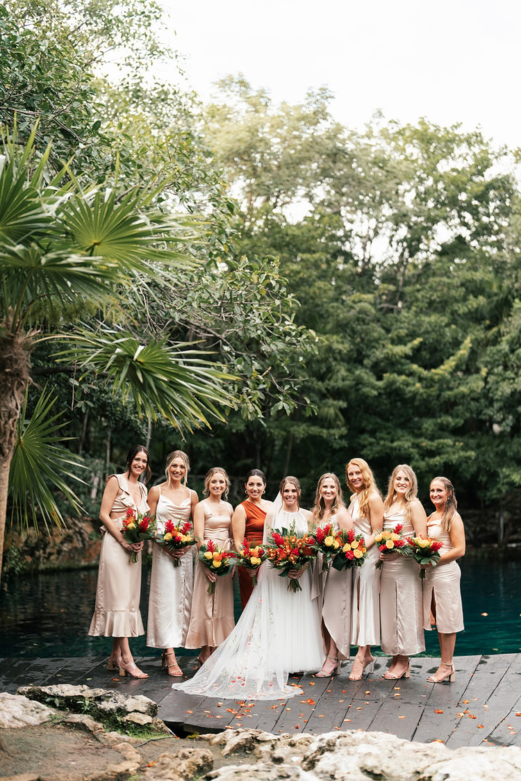 Bride poses with her bridesmaids in blush dresses on a dock beside a turquoise cenote, surrounded by palm trees.
