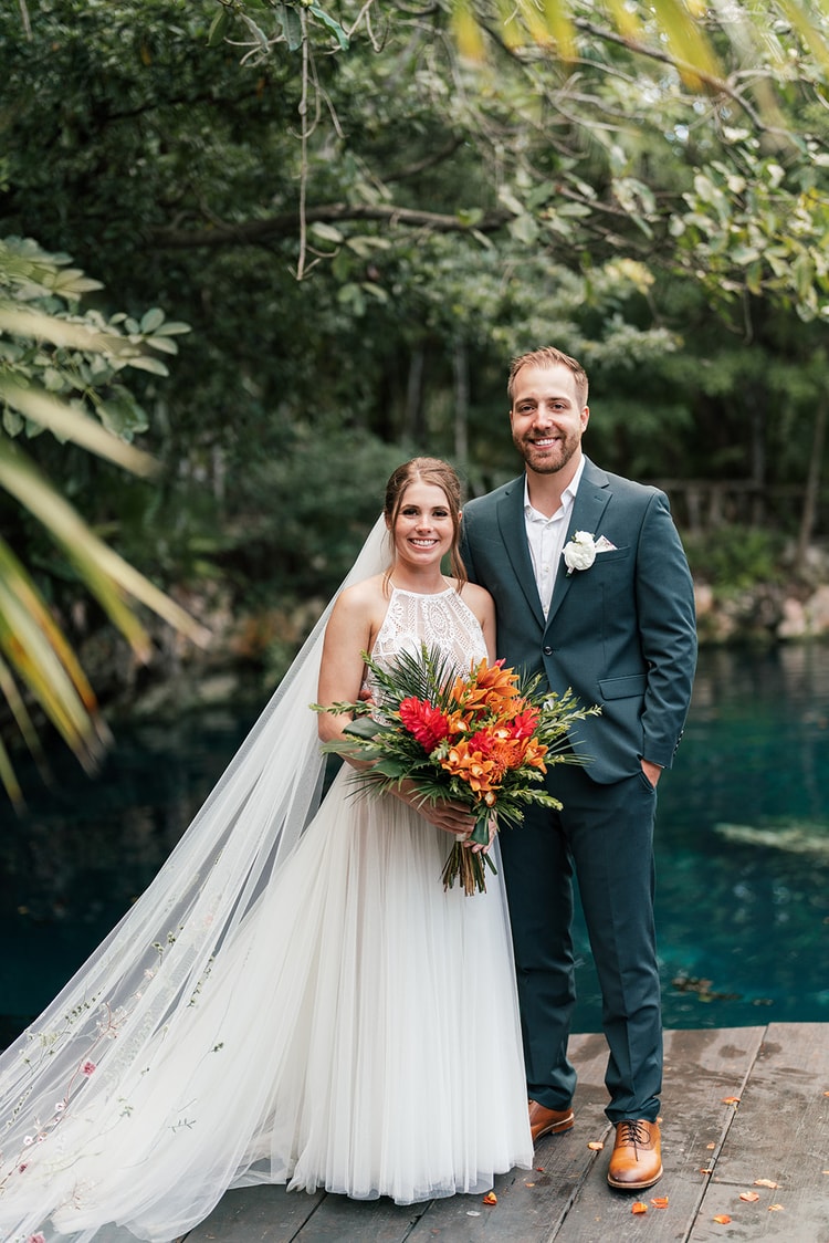 Bride and groom stand close together holding a tropical bouquet on a dock beside a jungle lagoon in soft daylight.