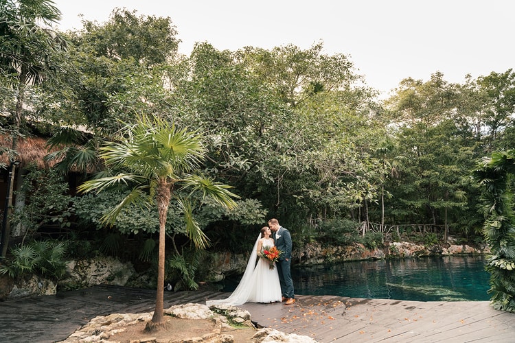 Bride and groom share a quiet moment beside a jungle cenote, a palm tree and turquoise water framing the wide scene.