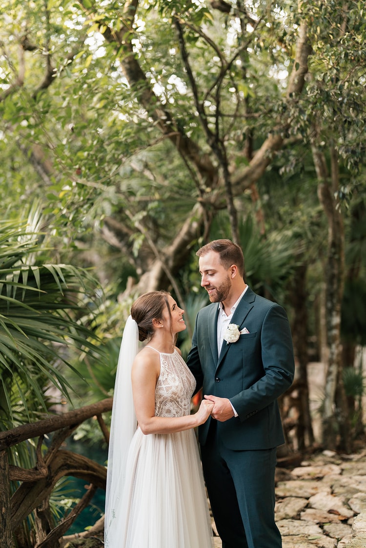 Bride and groom embrace closely on a stone path in dappled jungle light, her holding him gently by the shoulder.