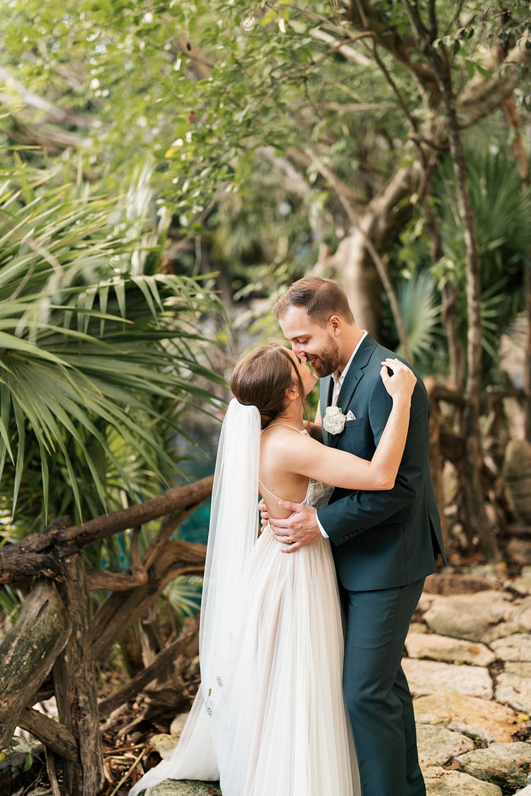 Bride and groom embrace and share a kiss beside a wooden railing next to a lush jungle cenote.