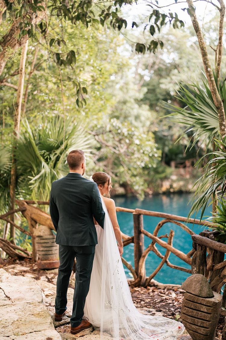 Bride and groom stand arm in arm gazing at a turquoise cenote from a rustic wooden railing, seen from behind.