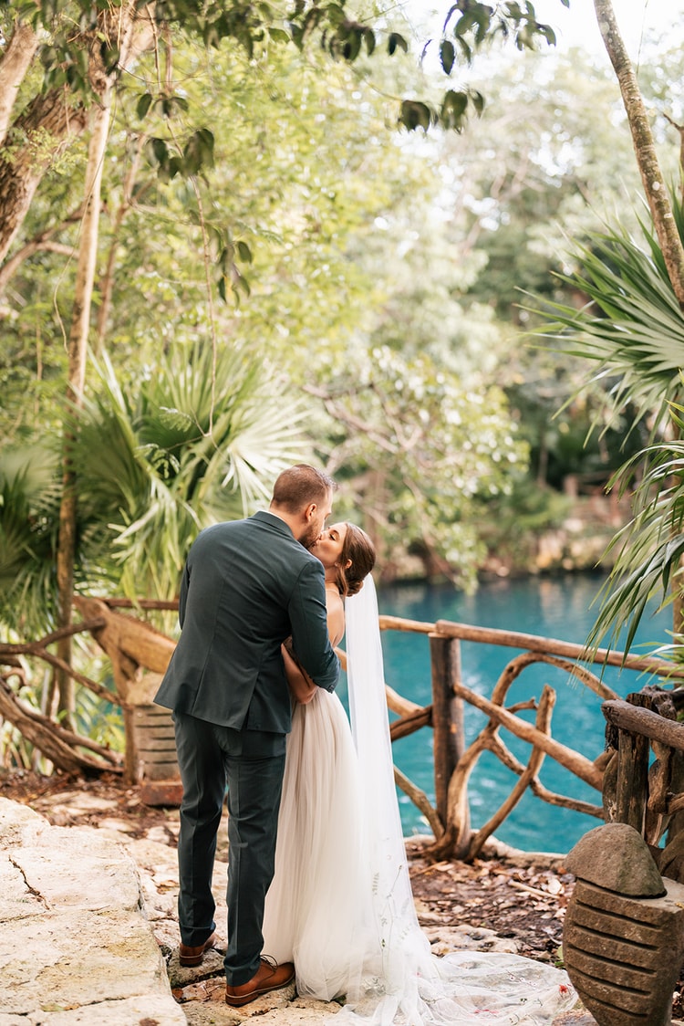 Bride and groom kiss beside a wooden railing overlooking a bright turquoise cenote, seen from behind among palm fronds.