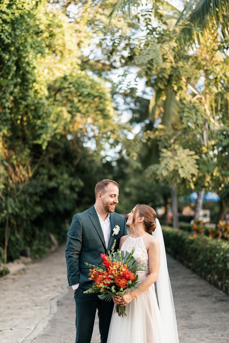 Bride and groom stand close together on a garden path, foreheads nearly touching, dappled sunlight filtering through palms.