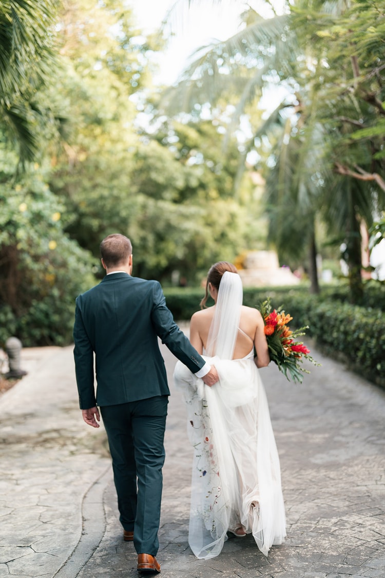 Bride and groom walk hand in hand away from the camera down a palm-lined path, her veil and bouquet trailing behind.