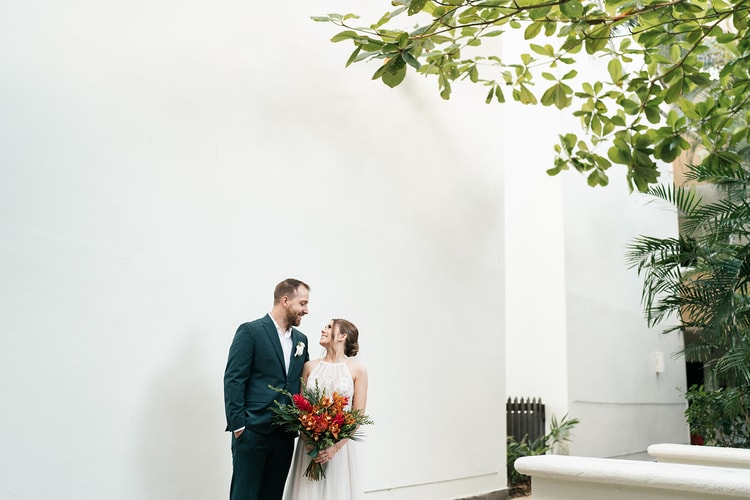 Bride and groom stand close together against a plain white wall, groom smiling down at her as she holds a bright bouquet.