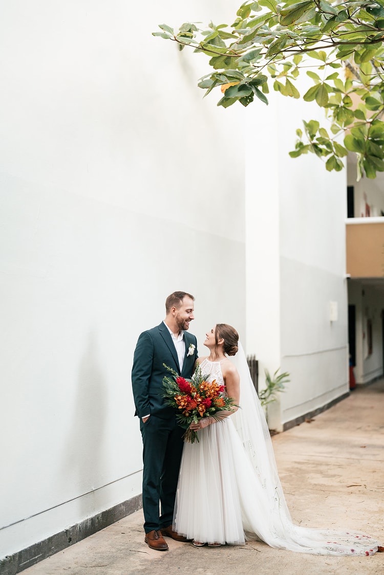 Bride and groom smile at each other beside a whitewashed wall in Playa del Carmen, her cathedral veil trailing on the ground.