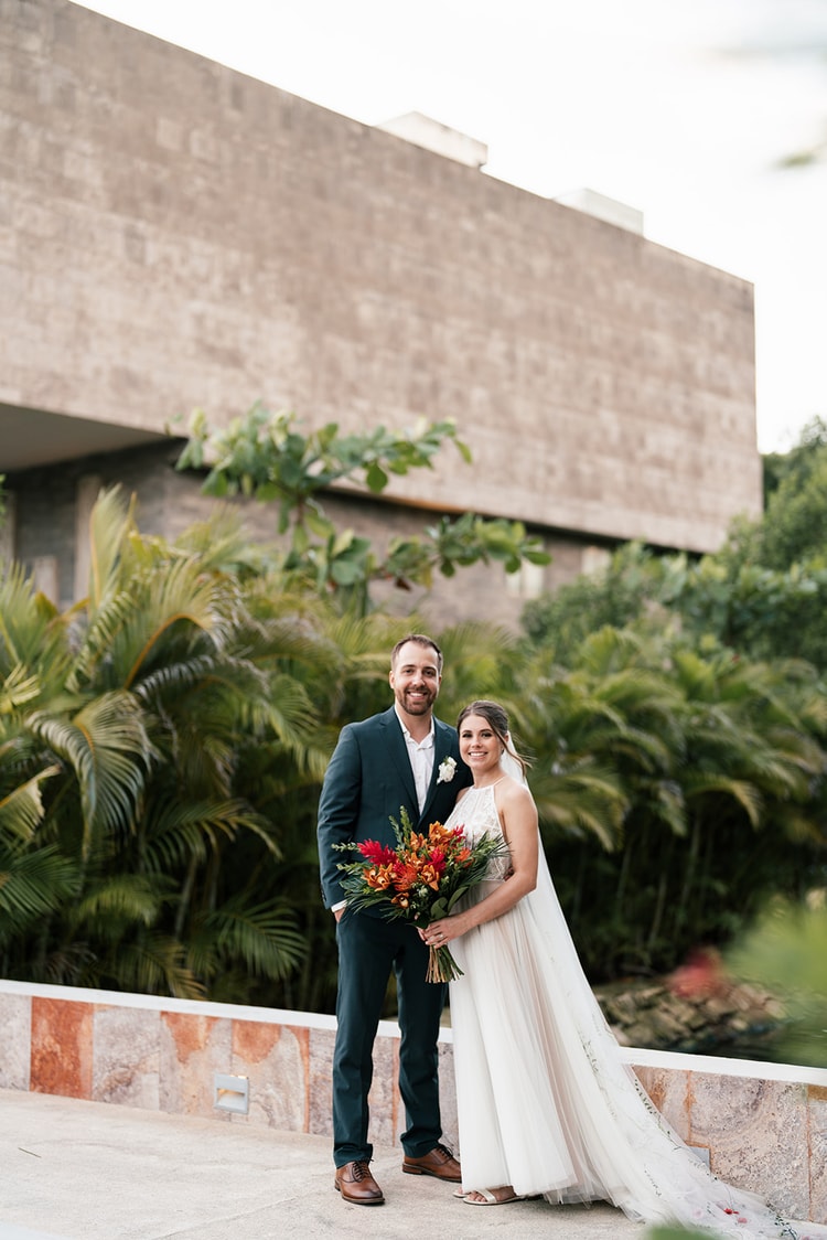 Bride and groom smile together on a terrace with a stone building and tall palms behind them at their Mexico resort wedding.