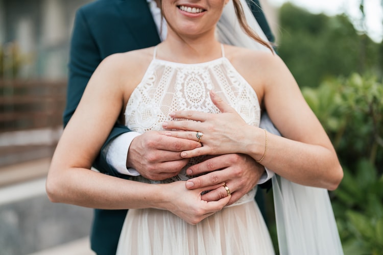 Close-up of the newlyweds' clasped hands showing an engagement ring and wedding bands against the bride's lace bodice.