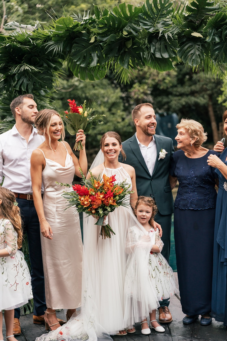 Bride and groom pose with family under a tropical leaf arch at their Playa del Carmen wedding, everyone smiling in soft daylight.