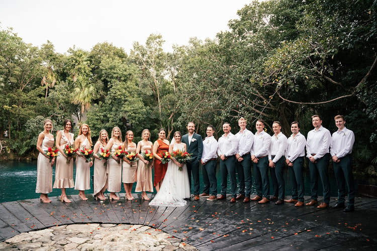Full wedding party lines a wooden dock beside a turquoise lagoon in Playa del Carmen, bride and groom centered in daylight.