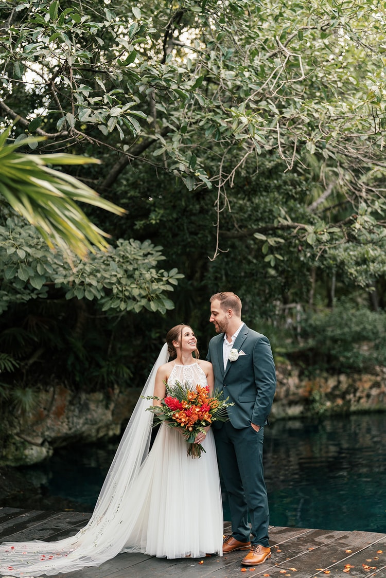 Bride and groom smile at each other on a wooden dock beside a turquoise lagoon framed by lush jungle greenery.