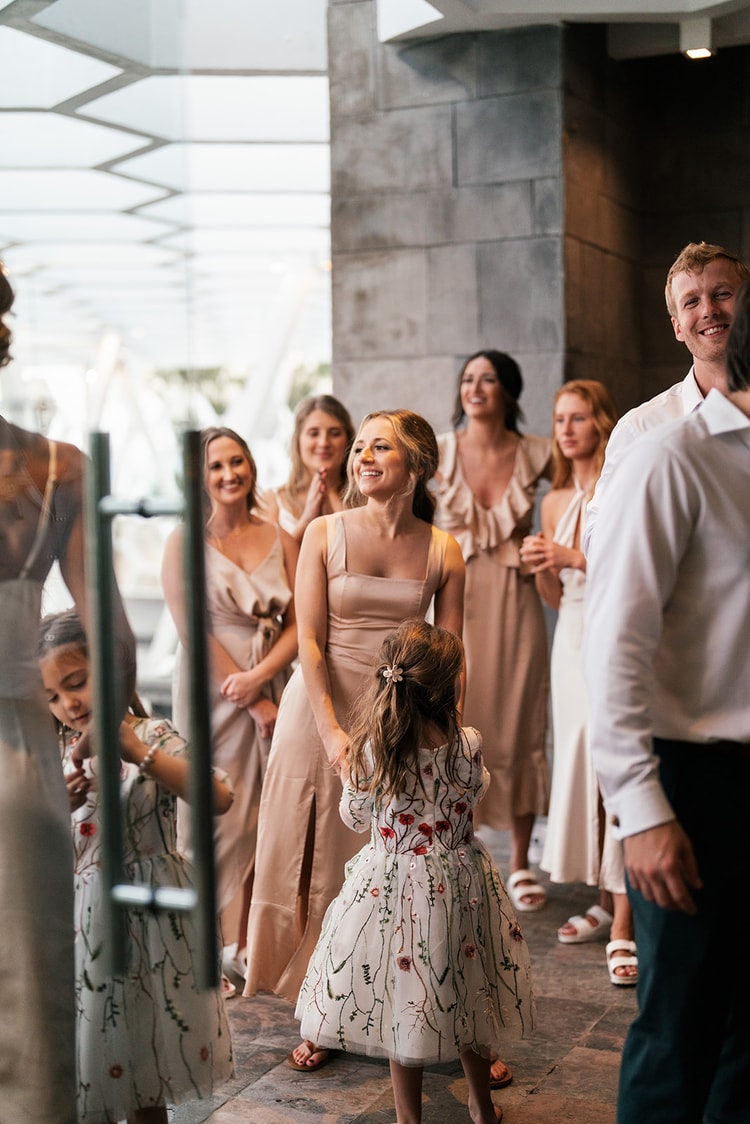 Bridesmaids and a young flower girl in an embroidered dress greet each other warmly at the indoor reception venue.
