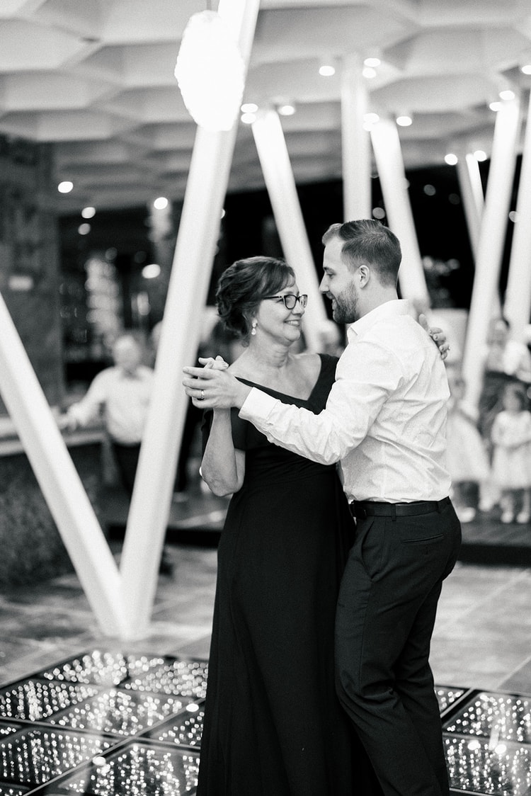 Groom shares a dance with his mother, who wears glasses and an off-shoulder black gown, on the reception dance floor at night.