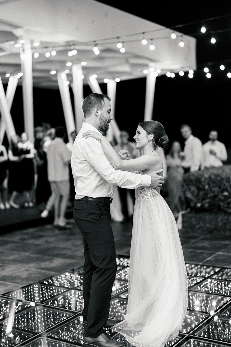 Bride and groom share their first dance, smiling at each other on an illuminated dance floor strung with string lights.
