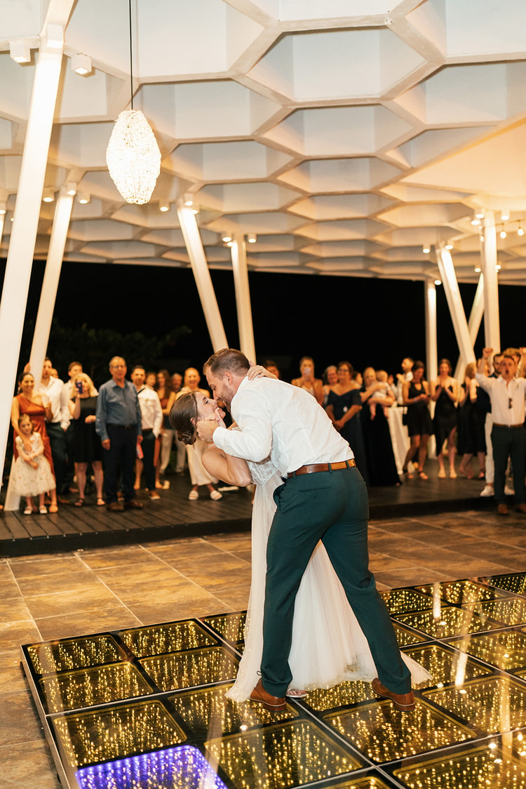 Groom dips the bride for a kiss on a lit-up dance floor as wedding guests cheer under a honeycomb-patterned pavilion ceiling.