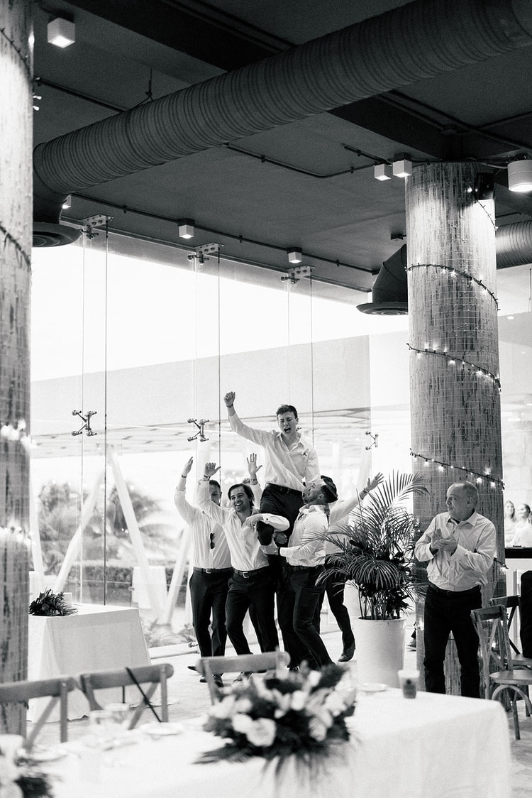 Black and white shot of groomsmen lifting a friend onto their shoulders in celebration inside the reception hall.