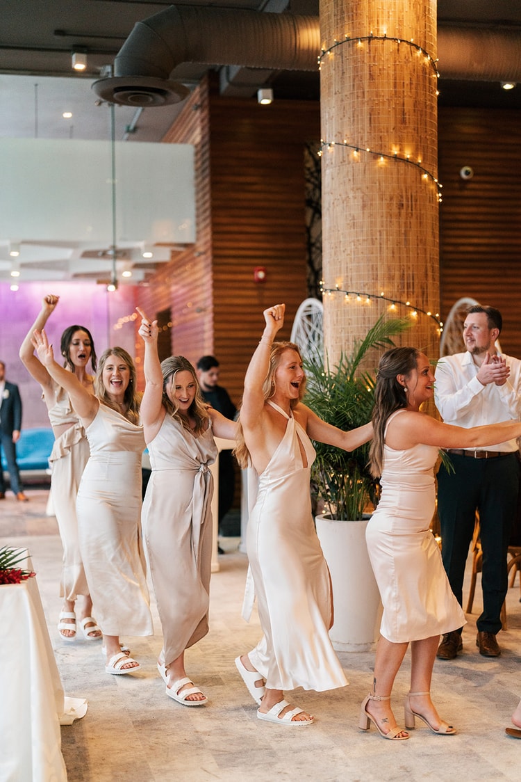Bridesmaids in satin dresses cheer and dance together at the reception, arms raised under string-lit wooden columns.